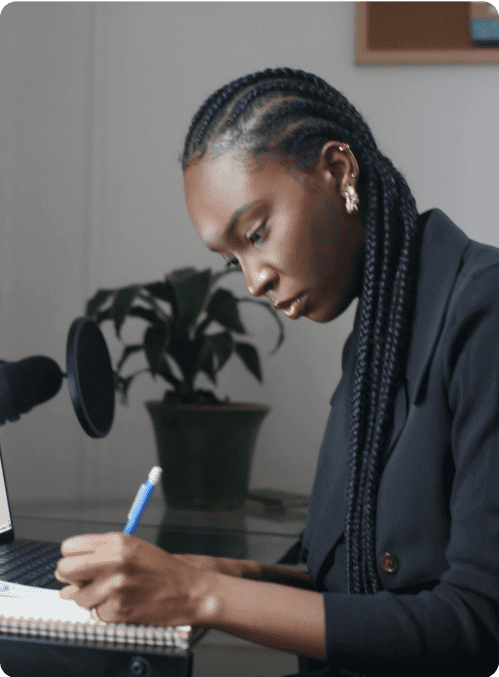 A woman, alone, writing in a notepad with a plant in the background.